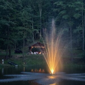 The EFL-250 Lighted Fountain sprays water on a pond at dusk, with a lit cabin and string lights among trees in the background.