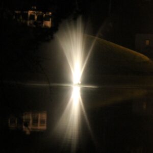 The EFL-190 Lighted Fountain glows at night, reflecting on a calm pond with distant buildings in the background.